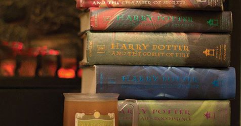 A mug of Butterbeer next to a pile of Harry Potter books with a fireplace in the background.