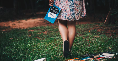 A woman wearing a dress walking away holding a copy of the book The Fault in Our Stars by her side.